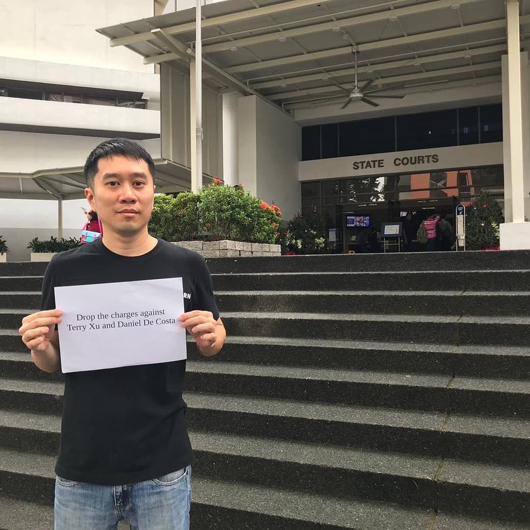 A Chinese Singaporean man in a black T-shirt and jeans standing in front of the State Courts. He is holding a sign printed on A4 paper that reads, “Drop the charges against Terry Xu and Daniel De Costa.”