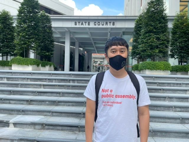A Chinese Singaporean man stands outside the State Courts. He is wearing a black face mask with a smiley face on it, and a white T-shirt that reads, “Not a public assembly. Just an individual in a t-shirt.”