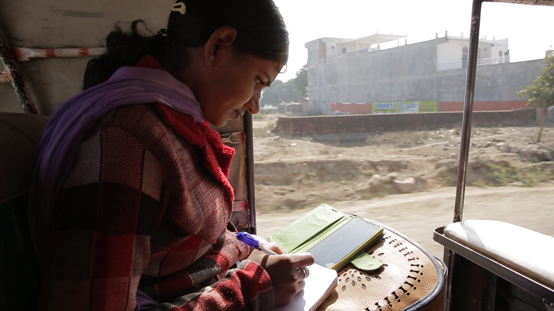 A young Indian woman in a red checkered top writes in a notebook using a purple pen. She is checking a smartphone for notes.