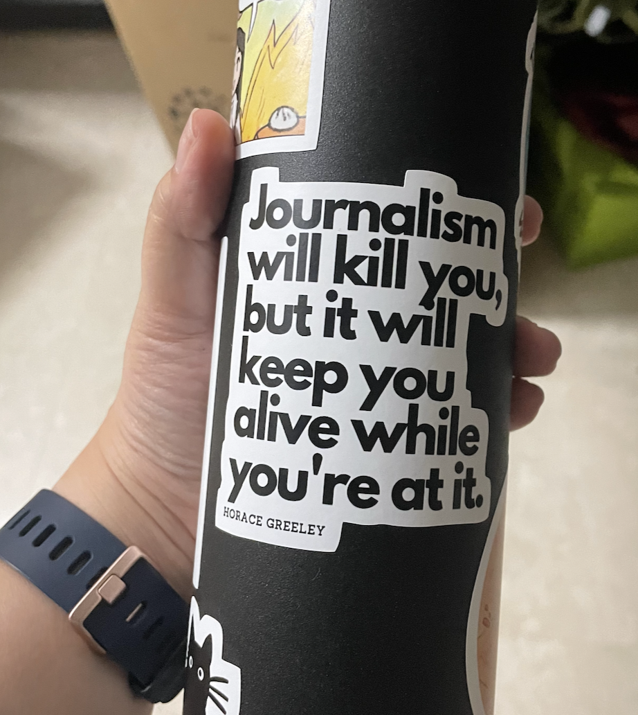 A white sticker with black text on a black tumbler. The sticker reads, "Journalism will kill you, but it will keep you alive while you're at it." It's a quote from Horace Greeley.