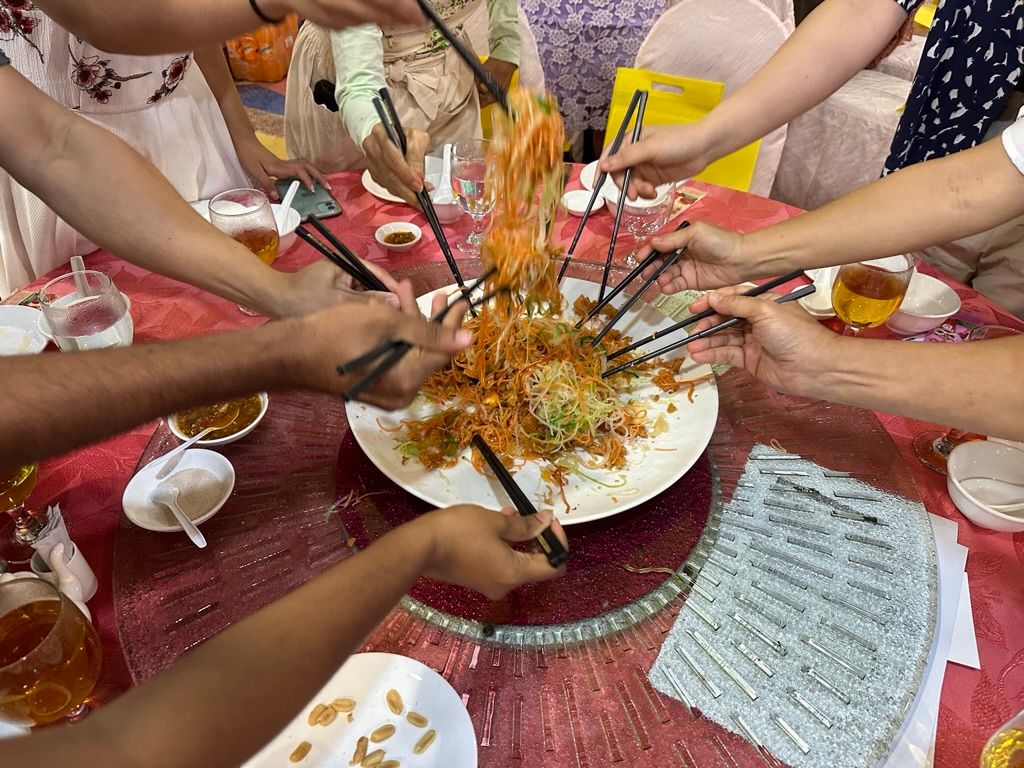 A photo of yusheng, on a glass lazy Susan, being tossed by hands holding black chop-sticks.