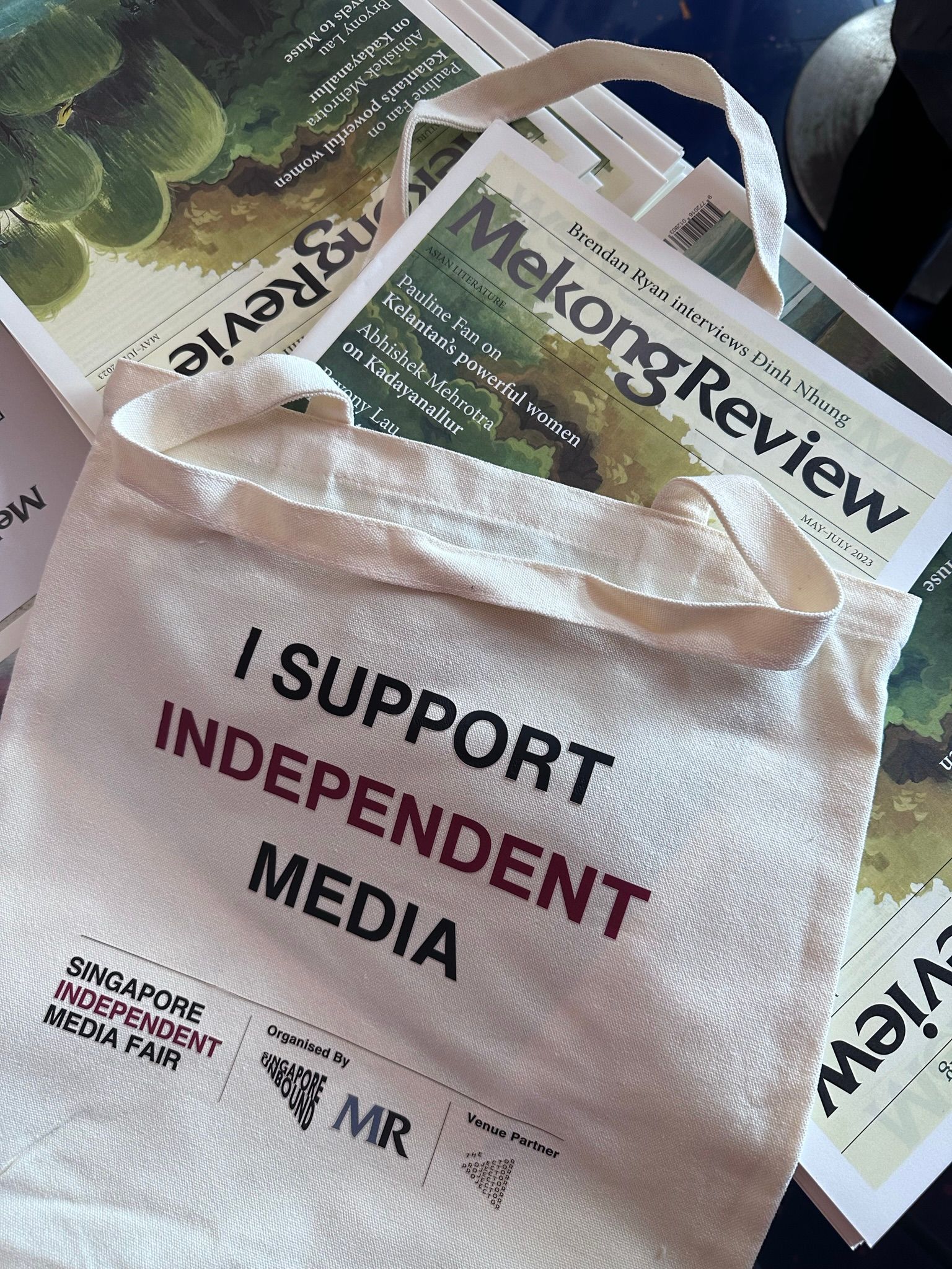 A photo of a canvas tote bag that says "I support independent media", with the Singapore Independent Media Fair branding at the bottom. There is a copy of the Mekong Review magazine half-inserted into it.